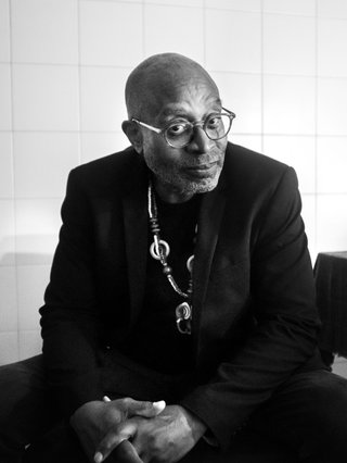 A black and white photo of Brian Jackson sitting down on a chair looking into the camera with his head slightly tilted and hands interlaced, wearing all black, glasses and a large necklace.