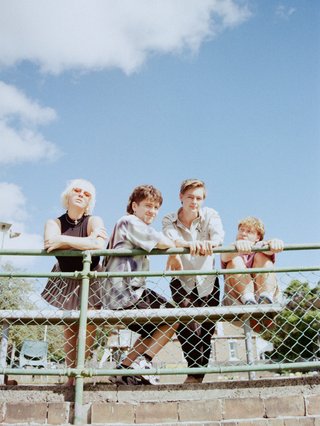 The four members of Faux Purr standing behind a metal fence before a light blue sky