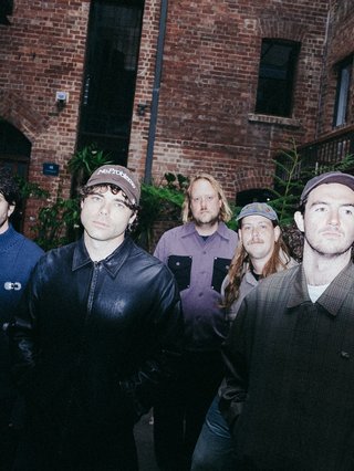 The five members of Naarm-based indie rock band The Velvet Club stand in front of a red brick wall and thick green ferns. They are wearing muted shades of blue denim, purple and neutrals.