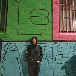 Amelia Sparke is wearing a black hoodie and pants, leaning against a blue, green and pink wall with drawings on it