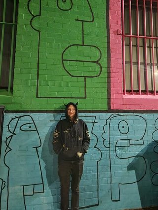 Amelia Sparke is wearing a black hoodie and pants, leaning against a blue, green and pink wall with drawings on it