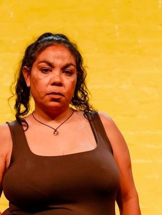 Angeline penrith stands stanuchly and tall in front of a brick wall painted with the Aboriginal Flag