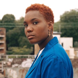 Arlo Parks stands in soft focus against a backdrop of construction and behind that green trees. She is turning her head to look at the camera with a calm expression on her face.