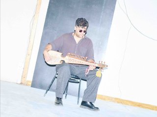Ashkan is sitting on a chair playing his rubab. He is wearing sunglasses, looking down in concentration. He is in a nondescript room and the image is overexposed stylistically.