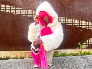 Attu crouches on gravel in front of a brown wall. They are wearing a white fur coat with a hood, holding a pink squid plushy. Attu has pink hair
