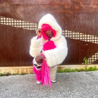 Attu crouches on gravel in front of a brown wall. They are wearing a white fur coat with a hood, holding a pink squid plushy. Attu has pink hair