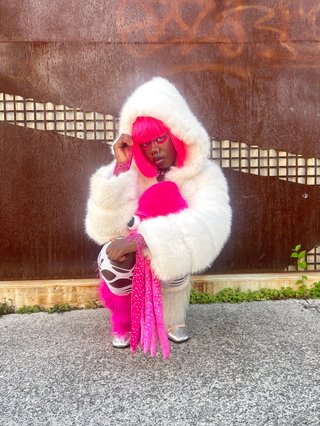 Attu crouches on gravel in front of a brown wall. They are wearing a white fur coat with a hood, holding a pink squid plushy. Attu has pink hair
