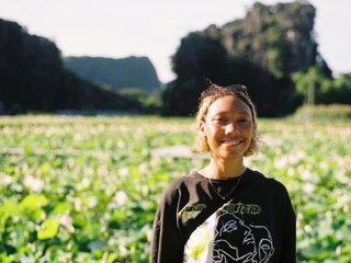 Bri is smiling and wearing a black t-shirt. She is standing in front of a lush, green field. There is warm light on her face from the left.