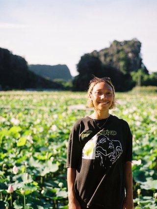 Bri is smiling and wearing a black t-shirt. She is standing in front of a lush, green field. There is warm light on her face from the left.