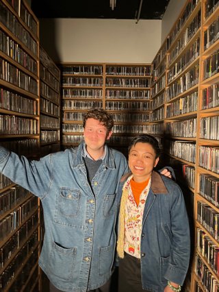 Harrie and Dr Xi Liu (Clinical Psychologist) stand in the music library in front of shelves filled with CDs - Both are wearing denim jackets and smiling into the camera