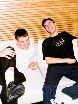 Both members of Chaos In The CBD in black and white clothing laying next to each other on a bed, smiling