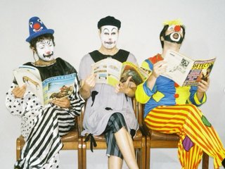 Three people dressed in clown outfits and mime facepaint sit on a bench against a white background. They are reading magazines