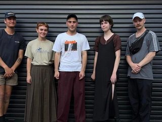 The five band members of Twine stand in front of a garage roller door in a line. They are all looking at the camera.