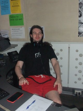 Jim Kretschmer is sitting in the studio staring into space appearing paralysed with their arms by their side and they are smiling.