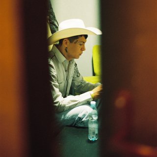 A side profile of Chuquimamani-Condori. They are wearing a cowboy hat and jeans, and a white cowboy hate. They are bathed in yellowish light. They are framed as if a crack in between a doorframe or two walls.