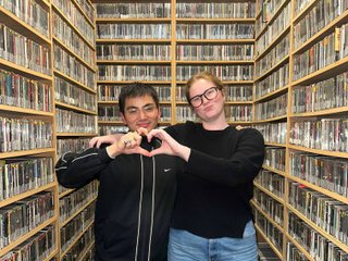 Matt and Kate stand in the music library and make a heart shape with one hand each.