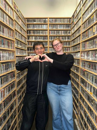 Matt and Kate stand in the music library and make a heart shape with one hand each.