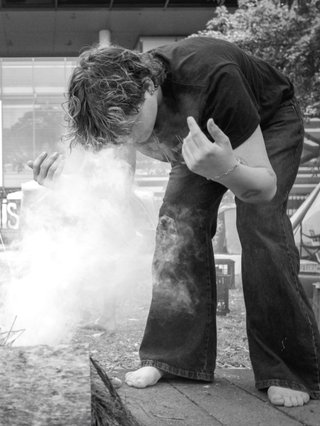 A black and white photo of Ethan Lyons. He's bending over ceremonial smoke and gesturing the smoke towards him