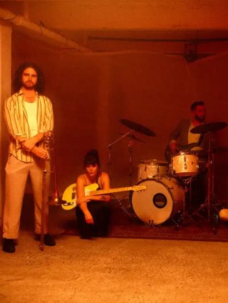 The five members of band FAIRTRADE NARCOTICS holding instruments around a drum kit in an orange lit room.