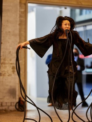 Image of artist WeiZen Ho during performance. She floating mid-air in a gallery and holding thick black ropes which also pour from her open mouth.