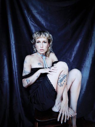 Freya Josephine Hollick. Blonde woman in black slip sitting in front of dark blue curtains