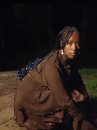 Fetle crouches in a park at night. They are wearing a long brown garment and looking solemnly into the camera