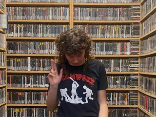 Maschinenkrieger stands in the fbi music library, looking down at the floor. she holds three fingers up on her right hand pointing up and three fingers down on the other hand pointing down