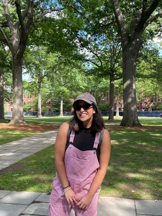 An image of a girl in white and red striped overalls with a high neck black top, sunglasses and a cap on, smiling in the sun. The backdrop is a park, with grass, trees and dappled light.
