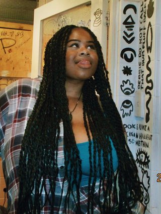 Ify - a Brown Skin girl with black braided twists, looking up to the side with her tongue sticking out. She wears a flannel and blue singlet in front of a graffiti wall background.