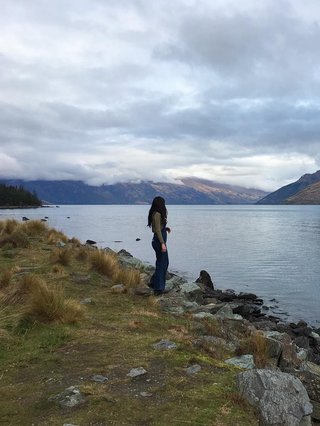 Hannah standing at the base of the lake in Queenstown, NZ. Back is turned, looking into the backdrop of clouds, mountains and water.