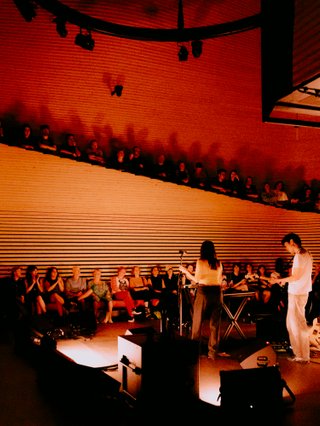 HTRK performing live at Phoenix Central Park, bathed in orange light. Viewed from the perspective of the corner of the venue, the band with their backs to the viewer, the crowd in view in the background.