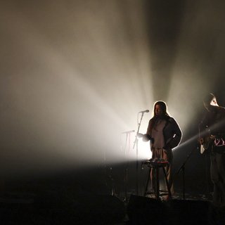 Image of HTRK on stage performing. The two members stand in the bottom right of the frame playing instruments. They are backlit by a white light, against a dark black background.