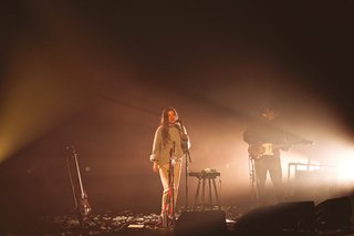 HTRK is performing lit by dusty warm white light. The signer is in the centre of the frame, singing into the mic. The guitarist stands in the background to the right, focusing on playing his instrument.
