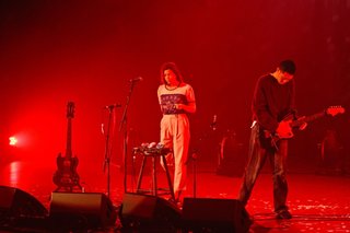 The two members of HTRK on stage at the Sydney Opera House, lit up by red lighting. One of them is playing a guitar, while the other is clicking in time to the music.