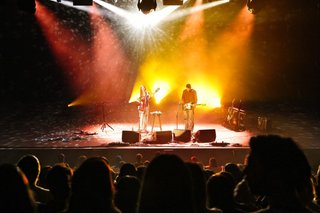 A shot of HTRK from the perspective of someone in the crowd. They are on stage playing their instruments lit up by orange and yellow light