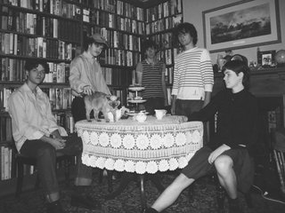 five members of Swapmeet assembled around a 20th century style table tea party setting. There is a cat walking across the table and it is being pet by one of the members. There is a fireplace and large bookshelves in the background.