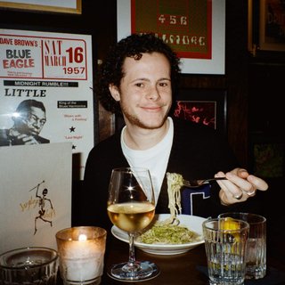 Sam blankly stares at the camera, brightly lit by a camera flash wearing a hat collared shirt and shoulder bag. Behind him a stage lit by the Aboriginal flag and a festival crowd.