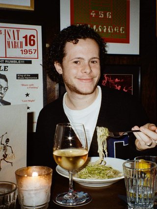 Sam blankly stares at the camera, brightly lit by a camera flash wearing a hat collared shirt and shoulder bag. Behind him a stage lit by the Aboriginal flag and a festival crowd.