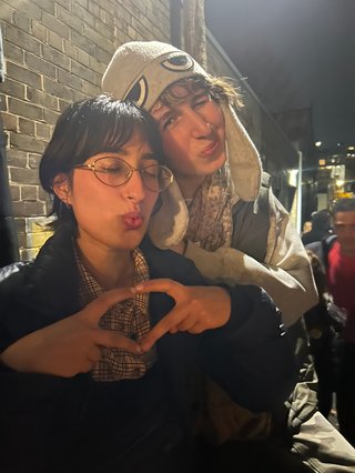 Two people sit outside smiling and leaning into each other. Their eyes are closed. One of them holds up their hands and fingers in a love shape