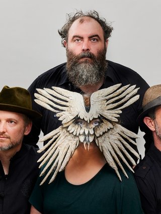 Album promo image for The Crooked Fiddle Band. Image has a white background, with Joe, Gordon and Mark wearing black and surrounding Jess, who is wearing a green shirt and a mask that covers her whole face and looks like four wings of a bird spread outwards.