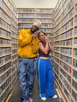 Sainté wearing a yellow jacket and a beanie, smiling with his hands next to his face. He is standing next to Bri who also has her hands next to her face in a spy pose.