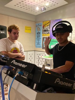 DAN+EE & FABZ stand behind the dj decks in the FBI studio. FABZ is mid mix, earphones on with one hand reaching for the headphones on his head. DAN+EE stands to the side, watching mid-groove, arms folded.