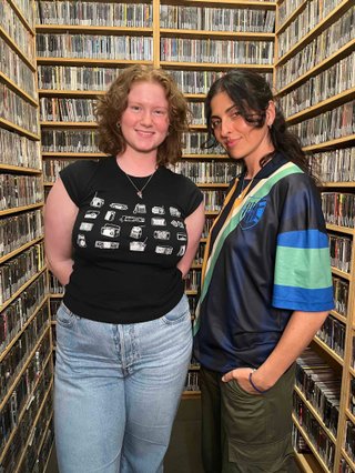 Kate and Anna pose in the music library. Kate wears a black t-shirt with white icons while Anna wears a blue, green and dark navy jersey.