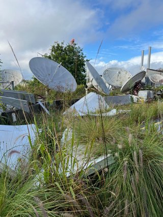 A collection of pale grey satellite dishes lying in tall grass against a pale cloudy blue sky