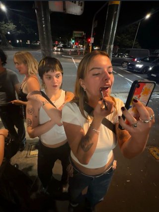 izzy is putting lip liner on looking at reflection in phone with friends behind her on the street