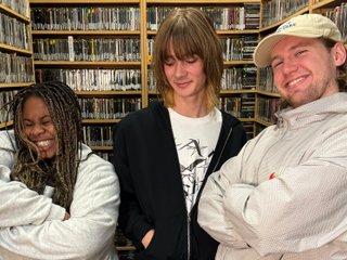 Up For It Hosts Ify and Benny and Independent Artist of the Week SLEEPR stand in the fbi.radio music library. They are surrounded by shelves of CDs. They are all posing for photo and smiling, Ify with her eyes scrunched closed, SLEEPR looking down and Benny leaning in and looking at the camera.