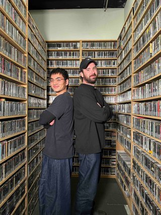 Matt and David stand cross armed back to back in the music library.