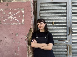 Jenna Parker is smiling at the camera, arms crossed in front of a worn down pink painted brick wall, and corrugated iron garage door. She has brown hair and bangs and is wearing a layered black shirt.