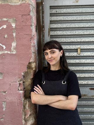 Jenna Parker is smiling at the camera, arms crossed in front of a worn down pink painted brick wall, and corrugated iron garage door. She has brown hair and bangs and is wearing a layered black shirt.