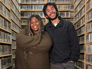 Up For It host Ify stands in the FBi Radio cd library with musician Jeromé Blaze. They are both facing the camera and smiling. Ify has her arms folded.